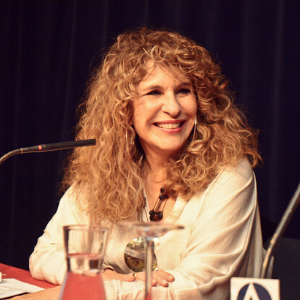 A woman with long, curly hair sits at a table, smiling, in front of a microphone. She is wearing a light-colored top and is seated against a dark curtain background. A glass of water and papers are in front of her.