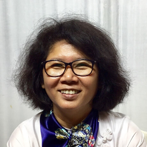 A woman with short, curly dark hair and glasses smiles at the camera. She is wearing a light-colored top and a patterned scarf. The background is a plain, light-colored curtain.