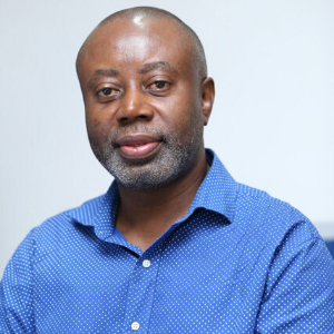 A middle-aged man with a short beard and shaved head, wearing a blue polka dot shirt, sits against a plain light background, looking into the camera with a neutral expression.