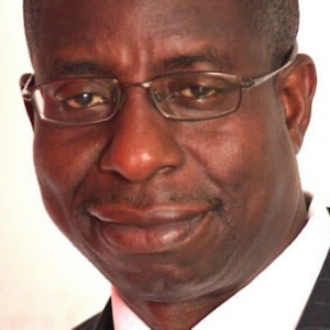 A middle-aged man wearing glasses and a suit, smiling softly at the camera against a plain background.