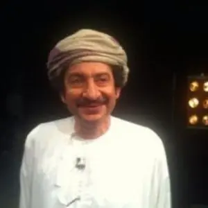 A man with a mustache wearing a traditional white robe and a wrapped headscarf stands smiling in front of a dark background with some lights visible on the right.