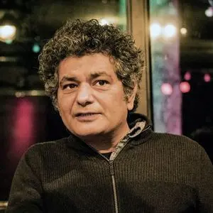 A man with curly gray hair, wearing a dark zip-up sweater, sits indoors with blurred colorful lights in the background.