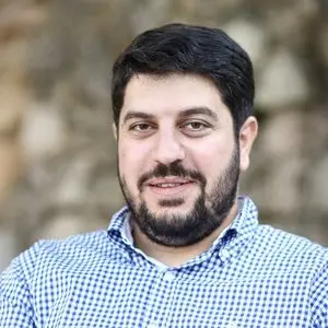 A man with dark hair and a beard, wearing a blue and white checkered shirt, smiles slightly while standing outdoors in front of a blurred stone background.