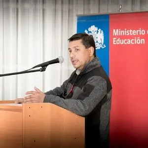 A man speaks at a podium with a microphone. Behind him is a red and blue banner with the logo and text for the Ministerio de Educación. White curtains are in the background.