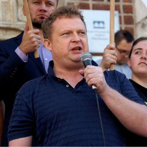 A man in a navy shirt speaks into a microphone at an outdoor event, with several people standing behind him holding wooden sticks.