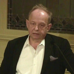 A man in a black jacket and white shirt, speaking at a podium indoors, with a patterned glass window in the background.