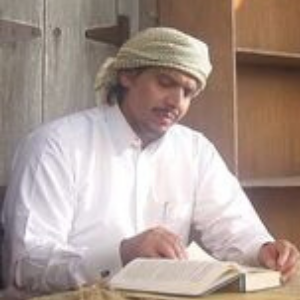 A man wearing a white thobe and a headscarf sits at a table reading a book, with shelves and wooden furniture in the background.