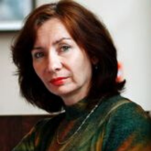 A woman with shoulder-length brown hair and light skin is wearing a green patterned top and a necklace, looking directly at the camera with a neutral expression.