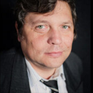 A man with light eyes and wavy brown hair wearing a dark suit, striped shirt, and loosely knotted tie looks into the camera against a dark background.