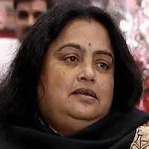 A middle-aged woman with long black hair and a small bindi on her forehead, wearing a black shawl, looks slightly to the side with a neutral expression. The background is blurred.