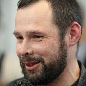 A man with a beard and mustache, wearing a dark jacket, smiles while speaking into a microphone. The background is blurred.