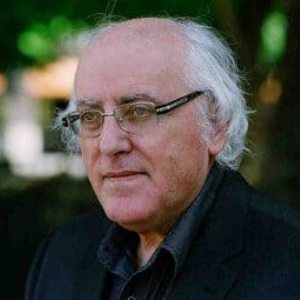 An older man with thinning white hair, glasses, and a black jacket stands outdoors with a blurred background of greenery and light.