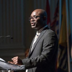 A man in a suit and glasses speaks at a podium with a microphone. Flags are visible in the background, and the scene appears to be at a formal event or conference.
