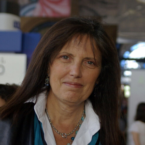 A woman with long brown hair and a white collared shirt stands indoors, looking at the camera with a slight smile. The background is blurred with people and signs visible.