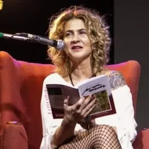 A woman with curly hair sits in a red chair, speaking into a microphone while holding an open book. She is wearing a white top and fishnet stockings, with part of a tattoo visible on her shoulder.