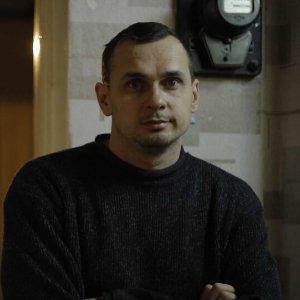 A man with short dark hair, wearing a dark sweater, stands indoors with his arms crossed. He is looking directly at the camera. There is a door and a black device on the wall behind him.