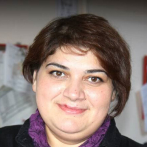 A person with short brown hair and a purple scarf smiling at the camera, with a blurred background featuring papers pinned to a wall.