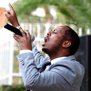 A man in a gray suit passionately sings into a microphone outdoors, with one hand raised and mouth open, against a blurred background of greenery and white fencing.