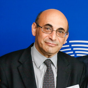 A middle-aged man with glasses, a bald head, and wearing a dark suit, light shirt, and patterned tie stands in front of a blue background with a partial white circular design.