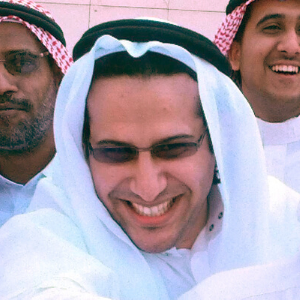 Three men wearing traditional Middle Eastern attire and glasses are smiling and laughing together, with one man in the center taking a selfie.