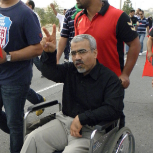 A man in a wheelchair wearing a black shirt and beige pants raises his hand in a peace sign while surrounded by people walking on a street.