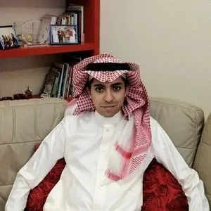 A man wearing a white thobe and a red and white checked keffiyeh sits on a beige couch with red pillows. Shelves with books, photos, and decorative items are in the background.