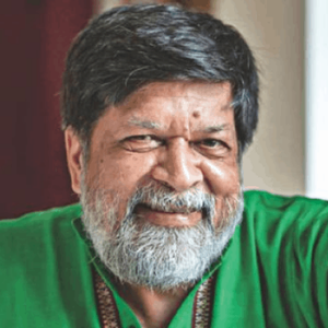 A middle-aged man with gray hair and a beard smiles warmly at the camera. He is wearing a green shirt and sitting indoors, with a blurred background behind him.