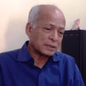 An older man with short gray hair, wearing a blue shirt, is sitting indoors in front of a beige wall and a black computer tower. He appears to be looking down pensively.