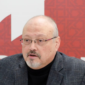 A middle-aged man with glasses, a closely trimmed beard, and a bald head is wearing a dark blazer and black turtleneck, sitting in front of a red and white patterned background.