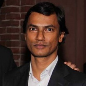 A man with short black hair wearing a dark suit jacket and light-colored collared shirt stands in front of a brick wall, looking at the camera with a neutral expression.