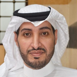 A man with a trimmed beard and mustache, wearing a white traditional Middle Eastern thobe and a white ghutra headdress with a black agal, poses indoors with a neutral expression.
