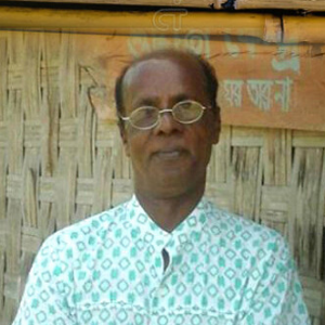 A middle-aged man wearing glasses and a patterned shirt stands in front of a woven bamboo wall with a wooden sign above him.