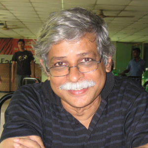A man with gray hair, glasses, and a mustache is smiling while seated indoors. He is wearing a black striped shirt. The background shows a cafeteria with other people and tables.