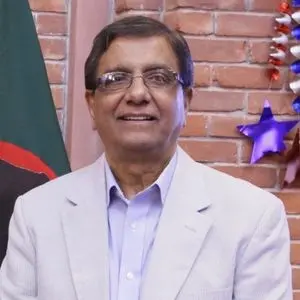 A man wearing glasses and a light-colored suit jacket smiles while standing in front of a brick wall decorated with stars and a flag.