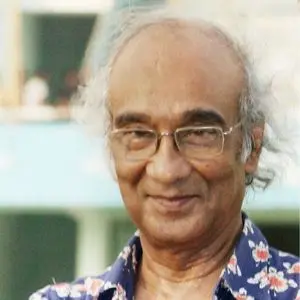 An elderly man with thinning gray hair and glasses smiles gently. He is wearing a dark shirt with a colorful floral pattern, and the background is softly blurred in blue and green tones.