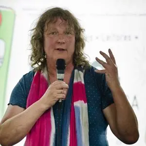 A person with wavy brown hair wearing a blue shirt and a colorful pink, white, and navy scarf speaks into a microphone, gesturing with their hand against a light-colored background.