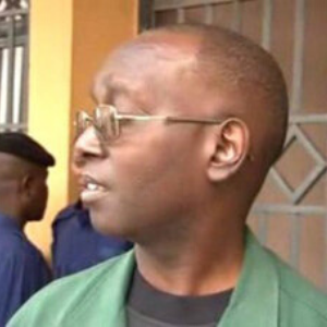 A person wearing glasses and a green jacket is shown in profile, looking to the left. Two other people in blue uniforms are visible in the blurred background near a metal gate.