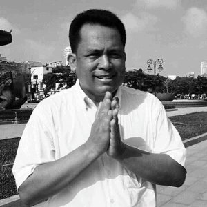 A man in a short-sleeved shirt stands outdoors, smiling and holding his hands together in a traditional greeting gesture, with buildings, trees, and a clear sky in the background. The image is in black and white.