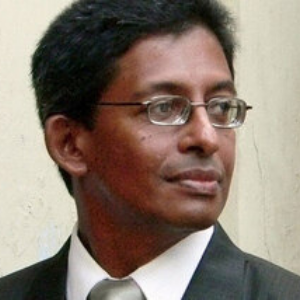 A man wearing glasses and a dark suit with a white shirt and tie looks to his right. He has short, dark hair and appears to be standing indoors near a light-colored wall.
