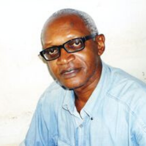 An older man with short gray hair and glasses is wearing a light blue button-up shirt. He is looking at the camera and sitting in front of a plain white background.