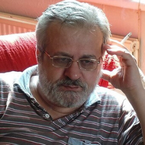 A middle-aged man with gray hair, glasses, and a beard sits on a red chair, wearing a striped shirt and holding a cigarette in his right hand, appearing deep in thought.