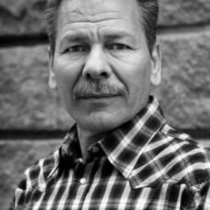 A man with a mustache wearing a checkered shirt stands outside, looking at the camera with a serious expression. The background features a stone wall. The image is in black and white.