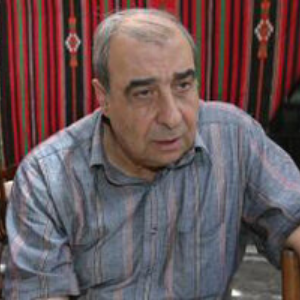 An older man with short gray hair wearing a striped shirt sits in front of a red and black patterned background.