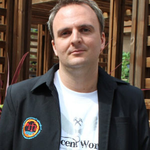 A man with short light brown hair wears a black jacket with a colorful patch and a white printed t-shirt, standing outdoors in front of wooden structures.