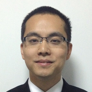 A man with short black hair and glasses, wearing a black suit, white shirt, and tie, smiles slightly while standing in front of a plain white background.