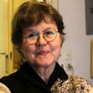 An older woman with short brown hair and glasses smiles softly. She is wearing a dark top and standing indoors, with a blurred light-colored wall and door visible in the background.