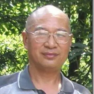 A middle-aged man with glasses and a shaved head stands outdoors in front of green foliage, wearing a gray collared shirt and looking at the camera.