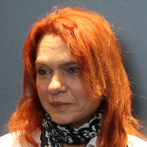 A woman with long, wavy red hair and fair skin is wearing a black-and-white patterned scarf and a light-colored top, posing in front of a plain gray background.