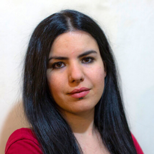 A woman with long straight black hair and fair skin is looking at the camera with a neutral expression. She is wearing a red top and is posed against a plain light-colored background.