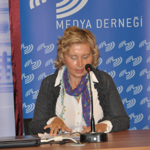 A woman with blonde hair speaks into a microphone at a table, with a laptop in front of her and a blue Medya Derneği banner in the background.